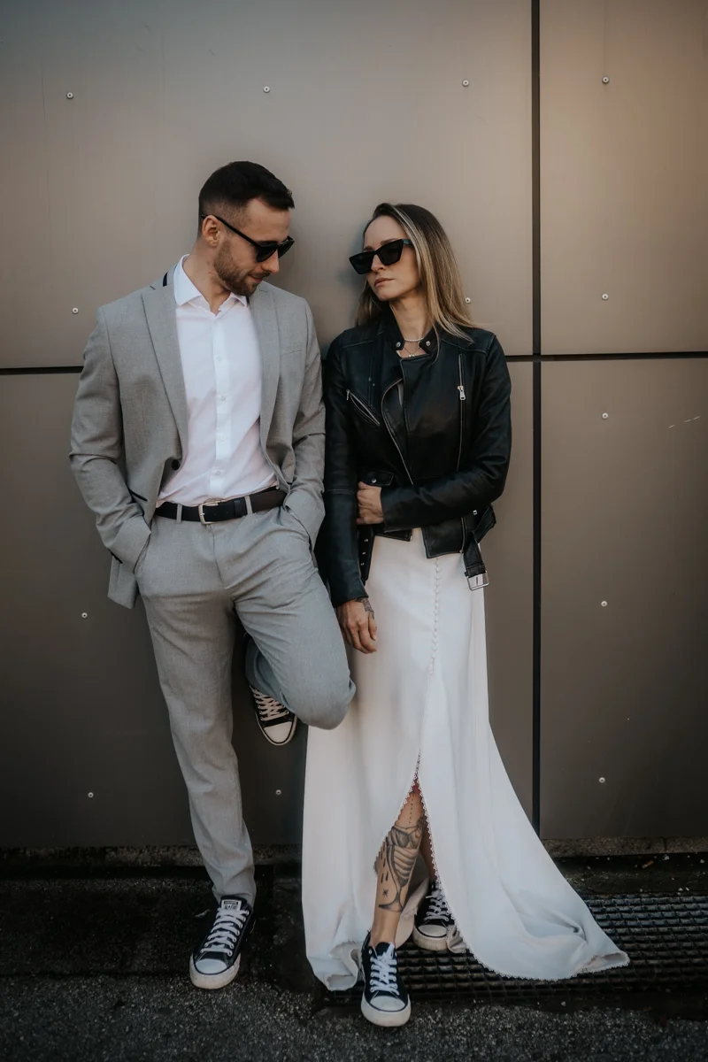 Cooles Brautpaar mit Sonnenbrillen, das sich gegen eine moderne Wand lehnt, fotografiert bei einem Hochzeit-Shooting in Wels