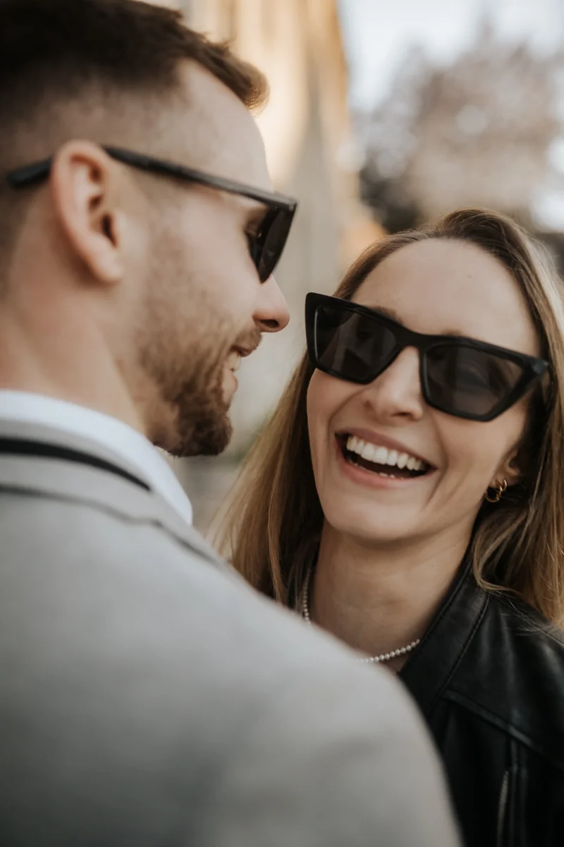 Lachende Frau mit Sonnenbrille, der Bräutigam trägt ebenfalls eine Sonnenbrille und lächelt seine Freundin an, fotografiert bei einem Hochzeit-Shooting in Wels