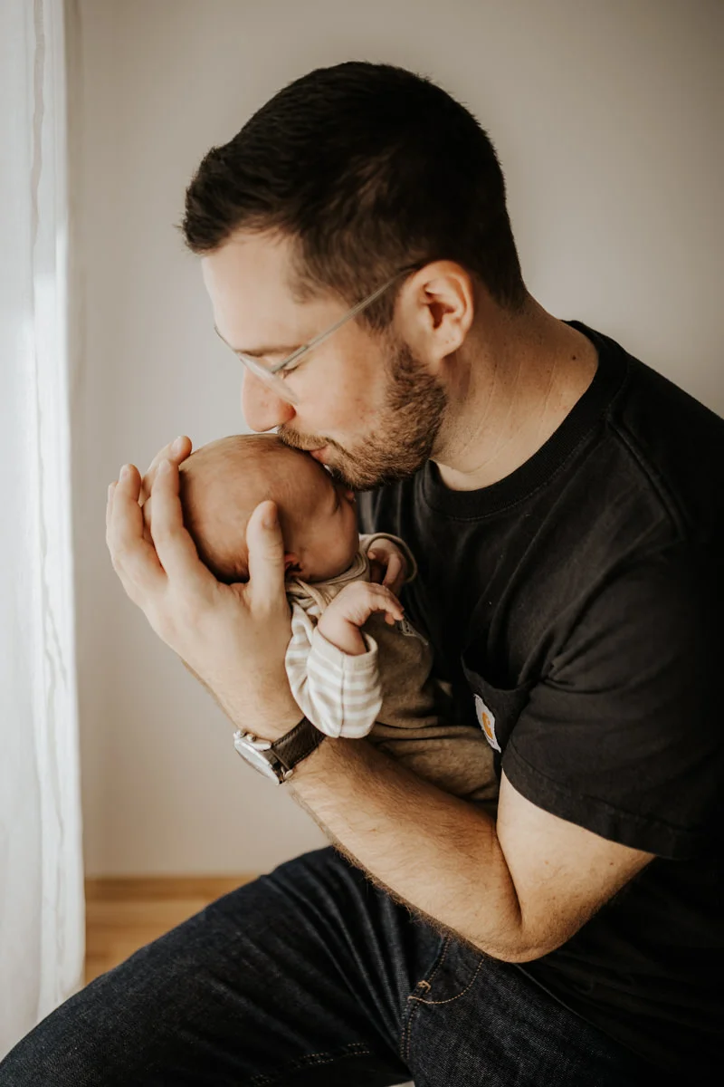 Vater, der sein neugeborenes Baby am Arm hält und küsst, fotografiert von Fotograf Kosia bei einem Newborn-Shooting in Oberösterreich