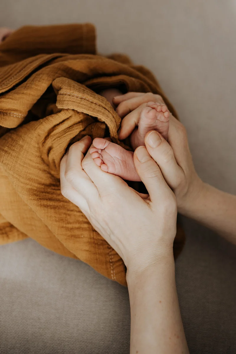 Detail-Foto von Füßen eines kleinen Neugeborenen, die in eine senf-farbene Decke gewickelt sind und von den Händen der Mutter gehalten werden, fotografiert von Fotograf Kosia bei einem Newborn-Shooting in Oberösterreich