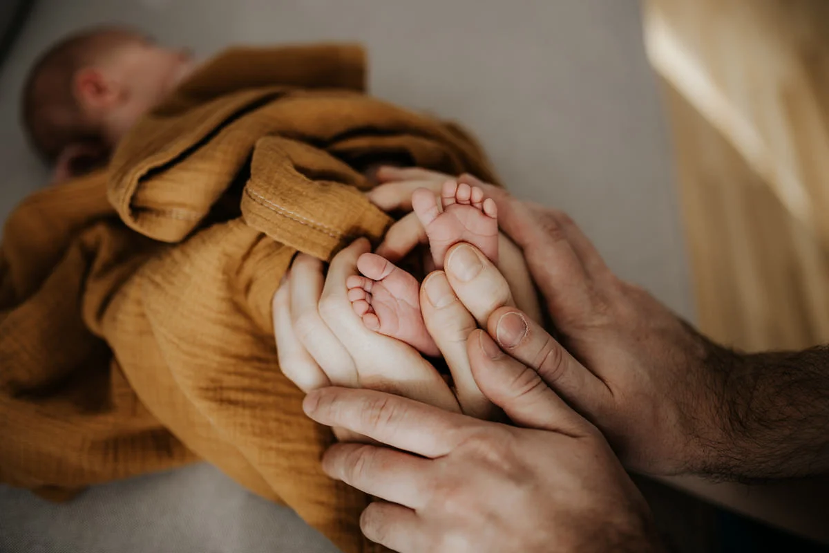 Detail-Foto von Füßen eines kleinen Neugeborenen, die in eine senf-farbene Decke gewickelt sind und von den Händen der Eltern gehalten werden, fotografiert von Fotograf Kosia bei einem Newborn-Shooting in Oberösterreich
