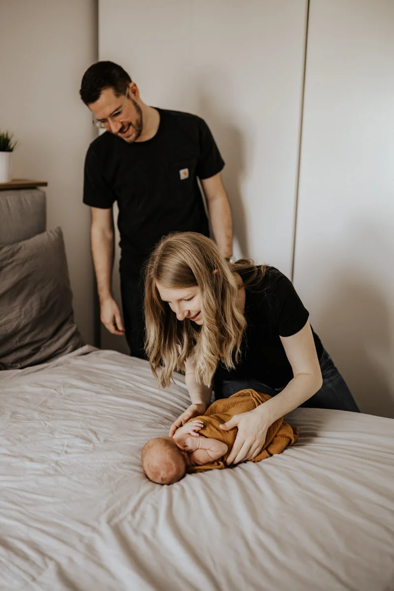 Junges Paar das im Schlafzimmer ihr neugeborenes Baby ins Bett legt, fotografiert von Fotograf Kosia bei einem Newborn-Shooting in Oberösterreich