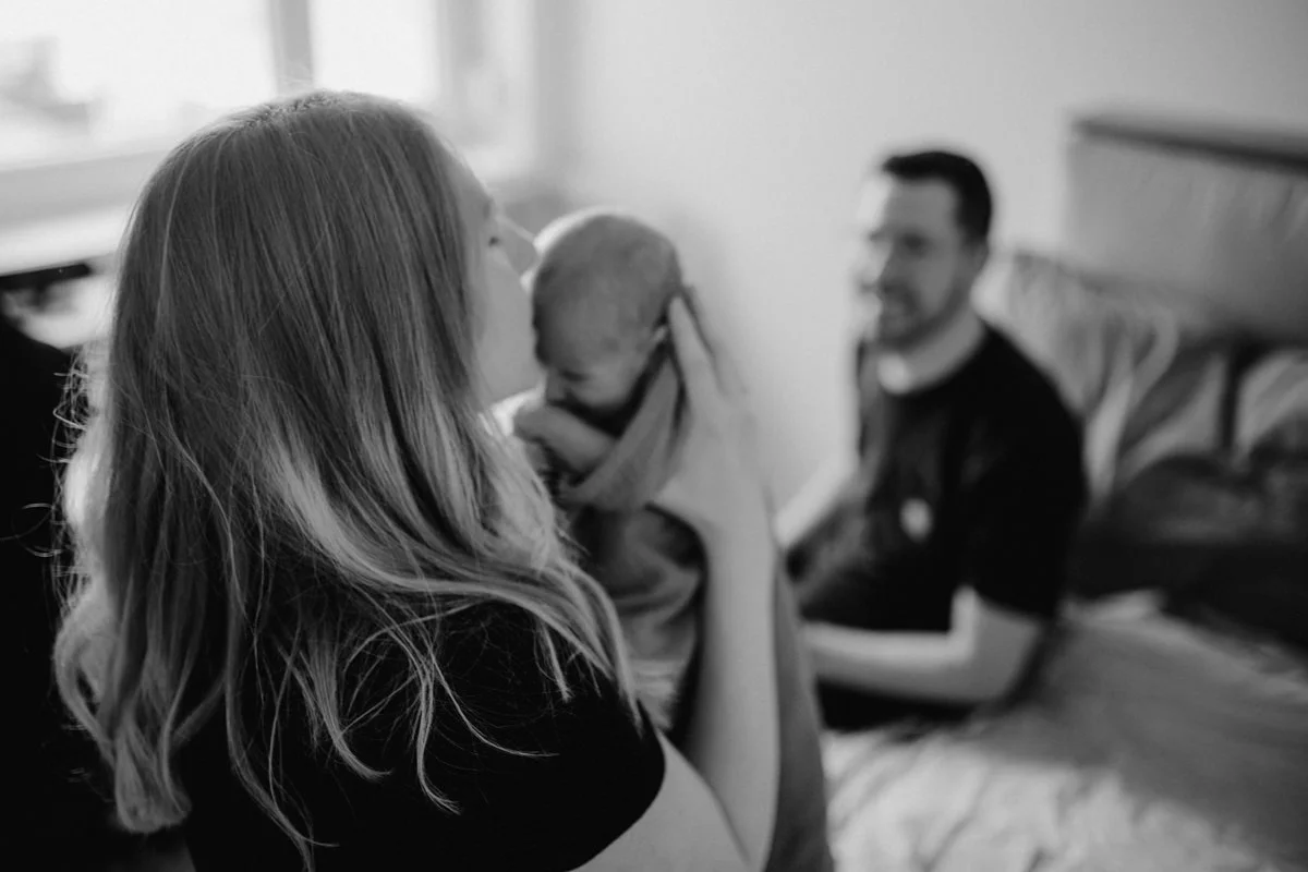 Schwarz-weiß Foto einer Mutter mit ihrem Neugeborenen Baby, mit dem Vater im Hintergrund, fotografiert von Fotograf Kosia bei einem Newborn-Shooting in Oberösterreich