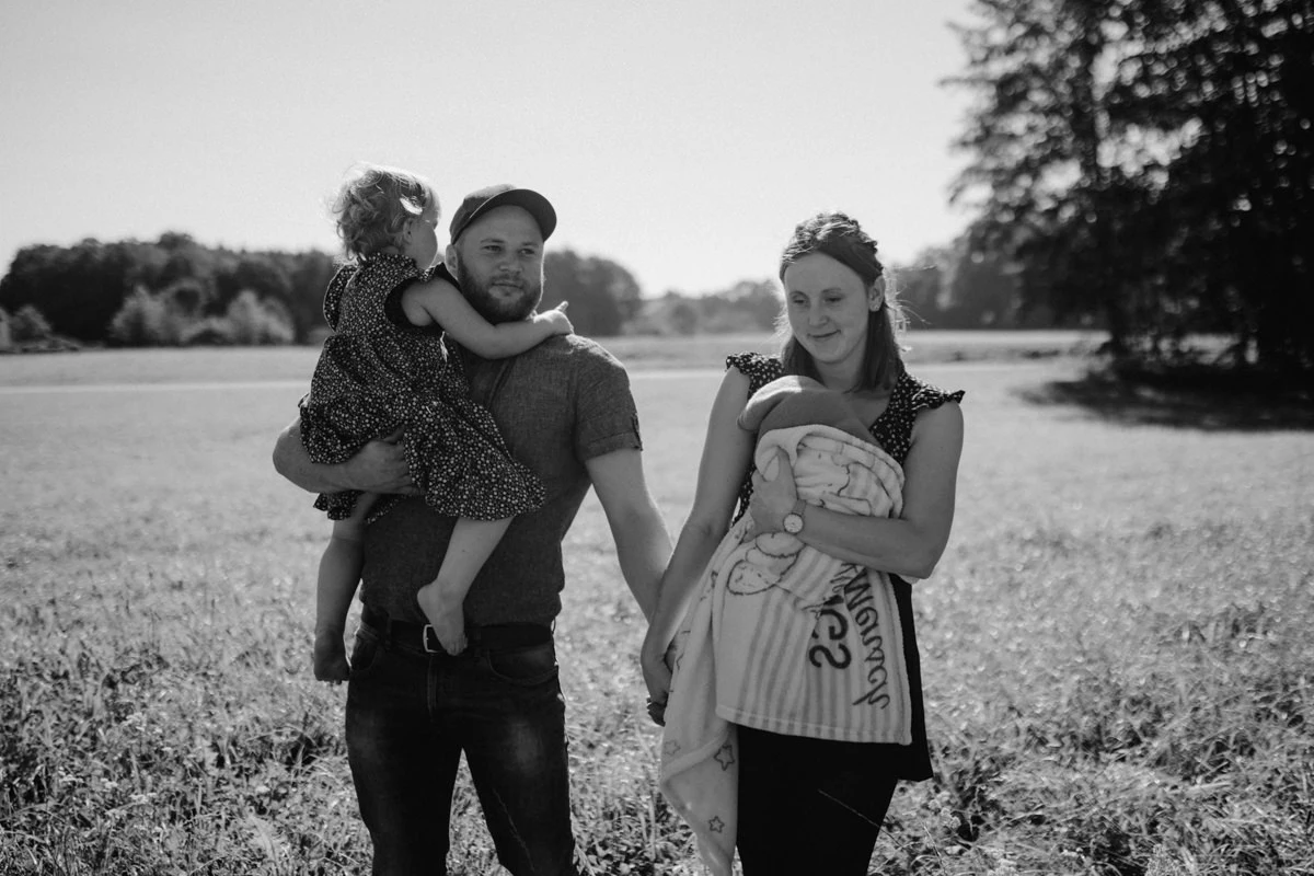 Eine Mutter mit Neugeborenem am Arm und ein Vater, der seine Tochter bei einem Newborn Shooting in Ried am Arm trägt, fotografiert von Fotograf Kosia