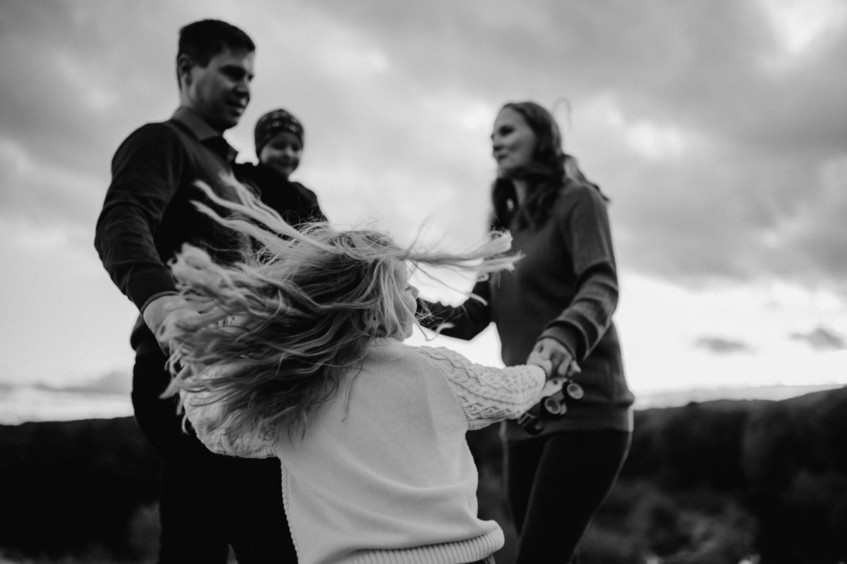 Schwarz-weiß Foto einer 4-köpfigen Familie, die gemeinsam im Kreis tanzt, fotografiert von Familienfotograf Kosia. Die Haare der Tochen fliegen im Wind. Authentische Familienfotos am Badesee im Bezirk Grieskirchen